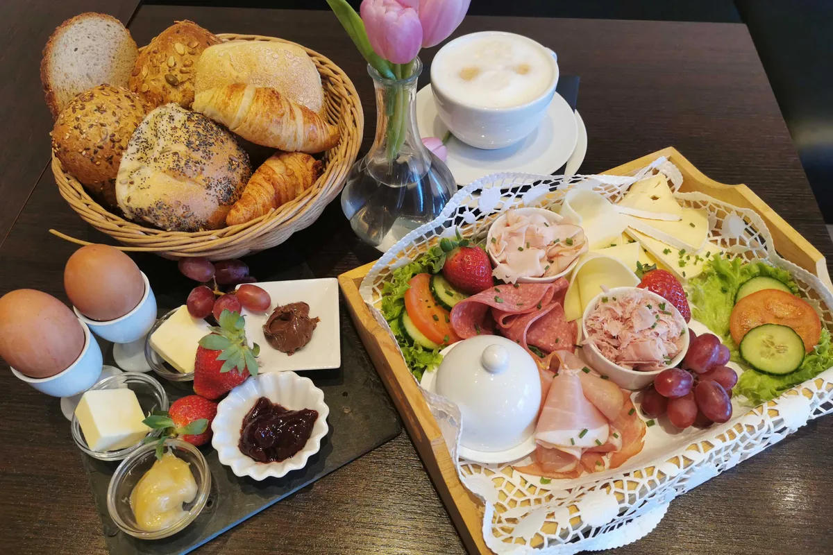Ein Frühstückstisch: ein Korb mit Brötchen und Croissants, gekochte Eier und eine Platte mit Aufschnitt, Käse und Obst.