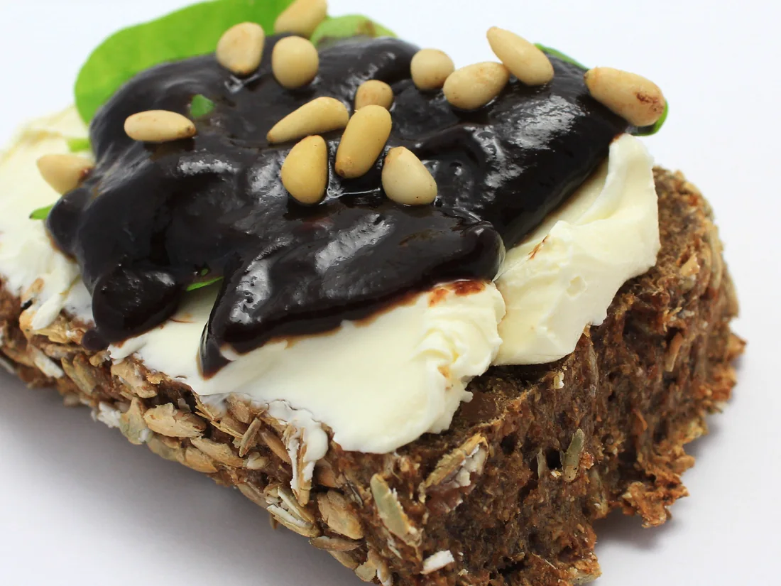 Eine Scheibe Schwarzbrot mit Frischkäse, Pflaumenmarmelade und Pinienkernen.