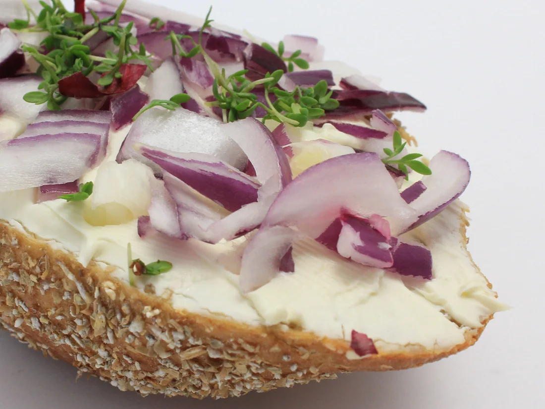 Eine Brötchenhälfte mit Körnerkruste, dick mit Frischkäse bestrichen, darauf rote Zwiebelringe und Kresse.