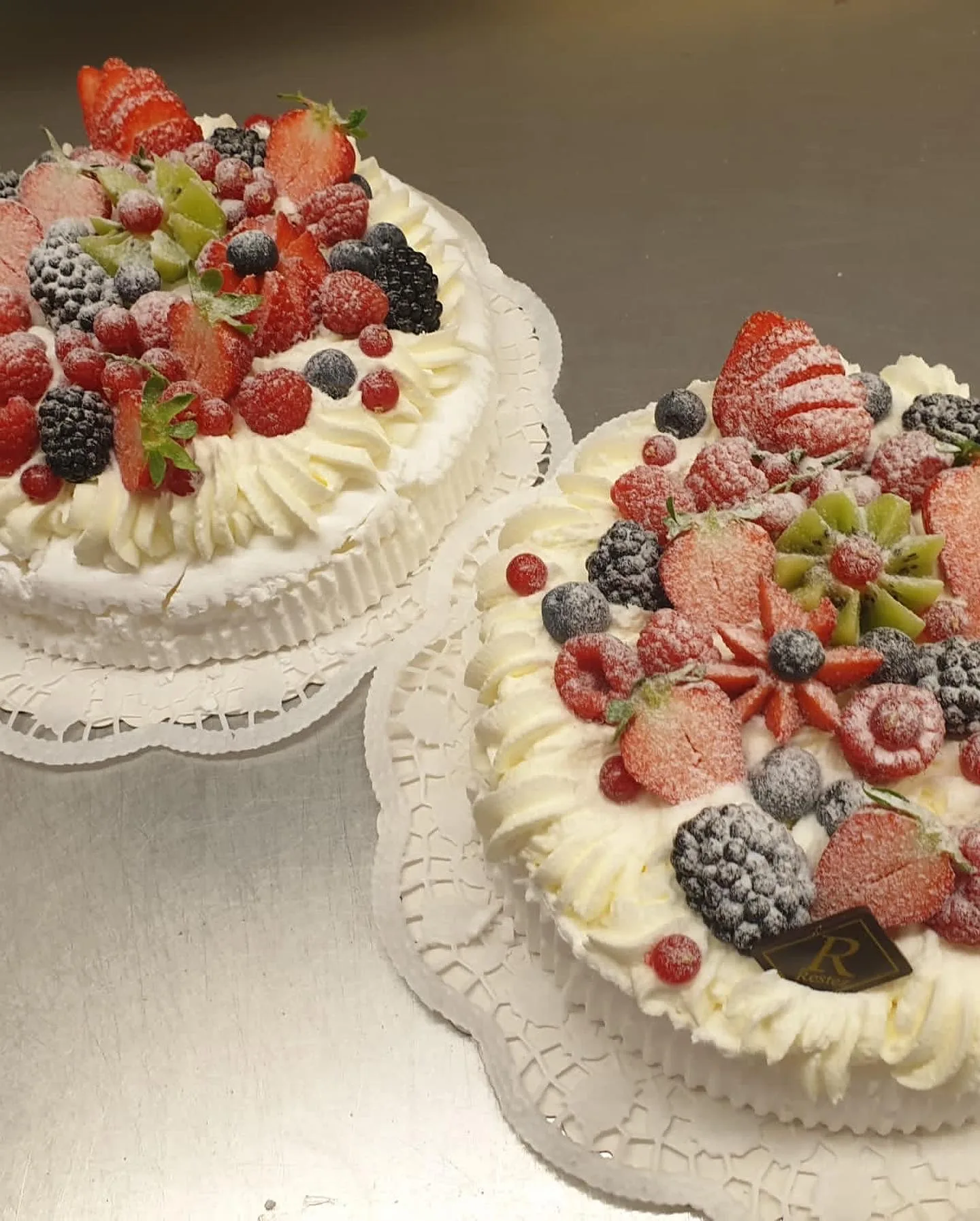 Zwei Sahnetorten, dicht belegt mit Erdbeeren, Himbeeren, Brombeeren und Kiwi, auf Tortenspitze.